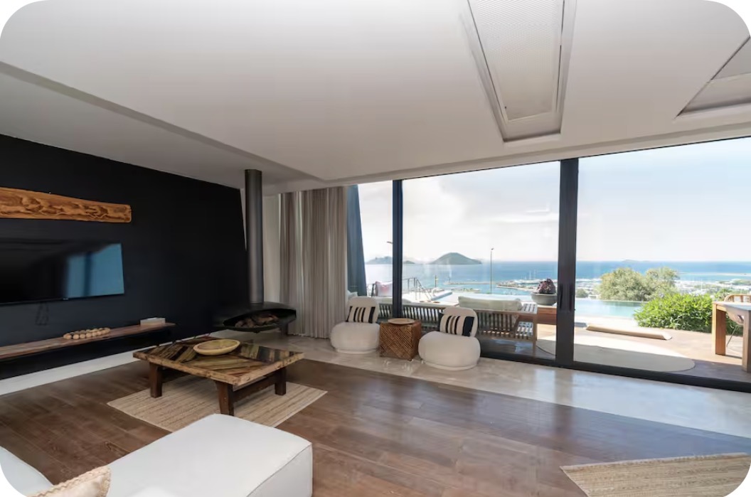 The villa's living room, with full-height glass onto the Bodrum bay and islands, a black stone fireplace wall and a solid wood table